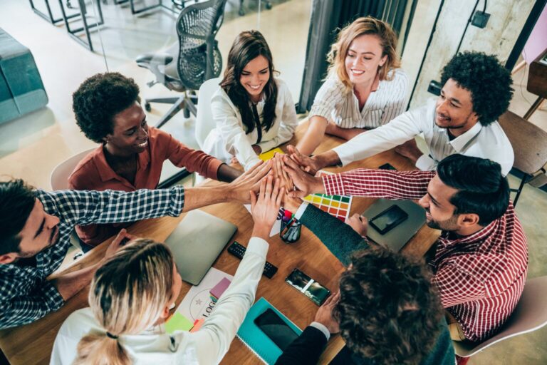 Equipe reunida ao redor de uma mesa juntando as mãos em gesto de união, representando como motivar sua equipe no dia a dia com ações simples.